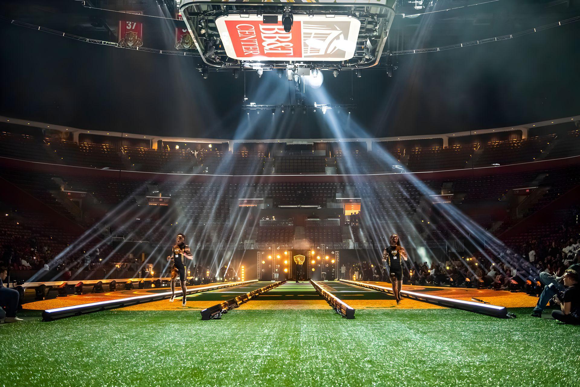 Indoor arena with spotlights and a runway, featuring athletes running on artificial turf.